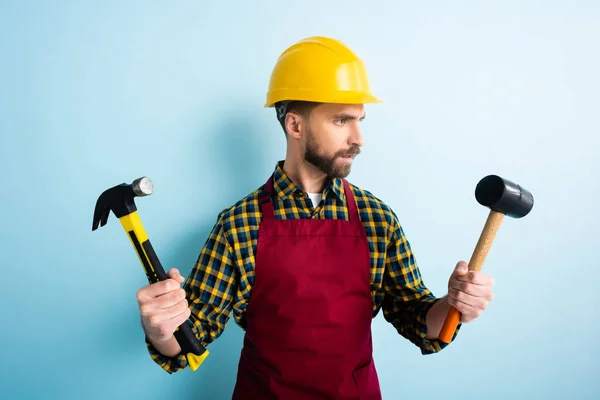 Handsome bearded workman holding hammers on blue — Stock Photo