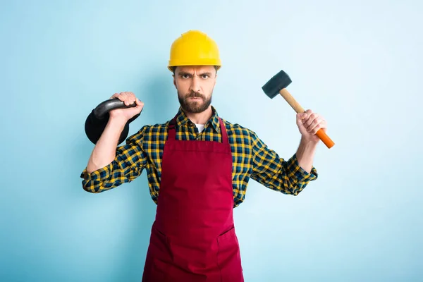 Злой бородатый рабочий держит молоток и гантель на синем — стоковое фото