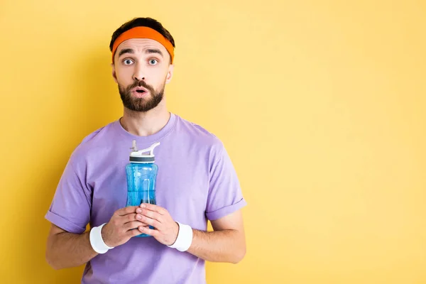 Desportista chocado segurando garrafa de esportes com água no amarelo — Fotografia de Stock