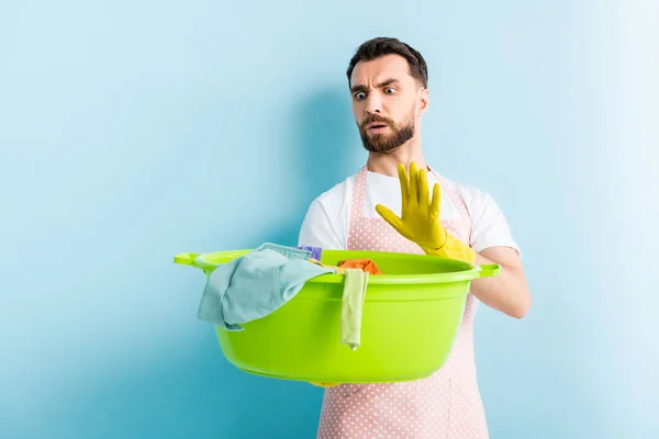 Schockierter Mann hält Plastikwaschschschüssel mit schmutziger Wäsche in der Hand und zeigt Stop-Geste auf blau — Stockfoto