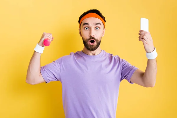 Sorprendido barbudo deportista sosteniendo pequeña mancuerna y teléfono inteligente en amarillo - foto de stock