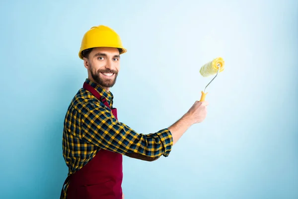 Cheerful worker in safety helmet holding paint roller on blue — Stock Photo