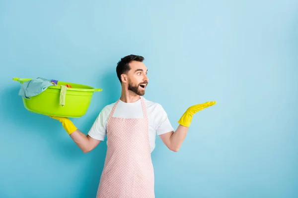 Hombre barbudo excitado sosteniendo la ropa sucia y señalando con la mano en azul - foto de stock