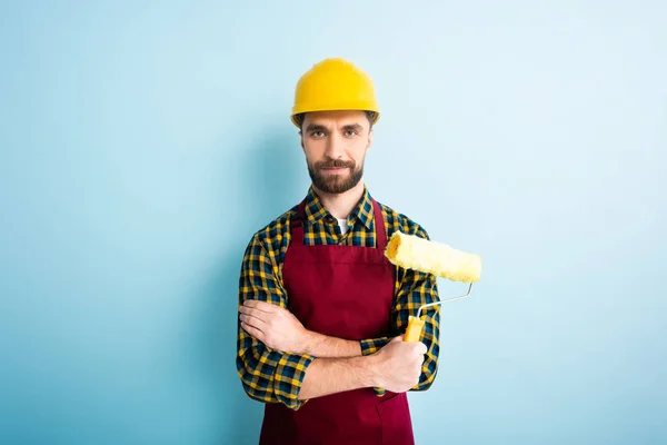 Positive worker in safety helmet holding paint roller on blue — Stock Photo