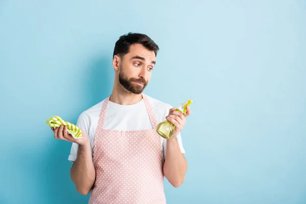 Bel homme barbu regardant chiffon tout en tenant bouteille de pulvérisation en plastique sur bleu — Photo de stock