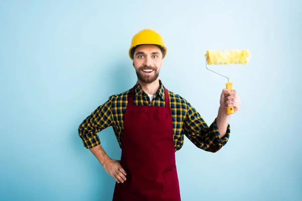 Positive worker in safety helmet holding paint roller and standing with hand on hip on blue — Stock Photo