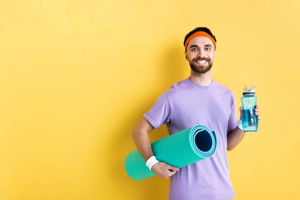 Positivo uomo barbuto in possesso di bottiglia sportiva e fitness mat su giallo — Foto stock