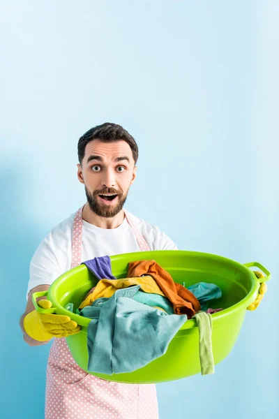 Шокированный мужчина, держащий пластиковую мойку с грязным бельем на голубом — стоковое фото