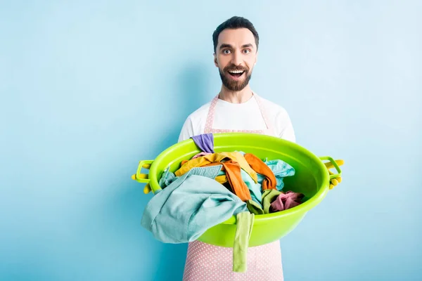 Cheerful bearded man holding dirty laundry in plastic wash bowl on blue — Stock Photo