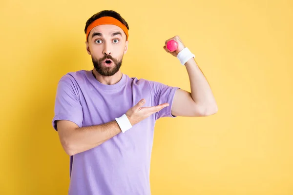 Surprised man pointing with hand at muscle while working out with small dumbbell on yellow — Stock Photo