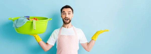 Plano panorámico de hombre barbudo excitado sosteniendo ropa sucia y señalando con la mano en azul - foto de stock