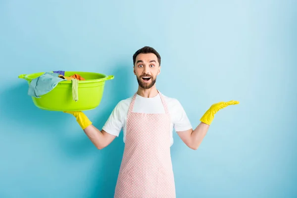 Hombre barbudo feliz sosteniendo la ropa sucia y señalando con la mano en azul - foto de stock