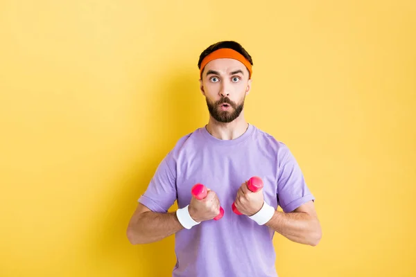 Shocked man looking at camera while holding small dumbbells on yellow — Stock Photo