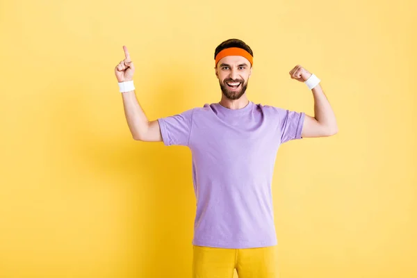 Cheerful sportsman pointing with finger while showing muscle on yellow — Stock Photo