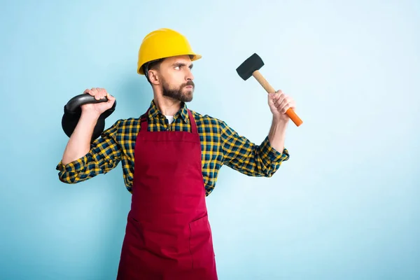 Wütender bärtiger Arbeiter mit Schutzhelm hält Hammer und Hantel auf blau — Stockfoto