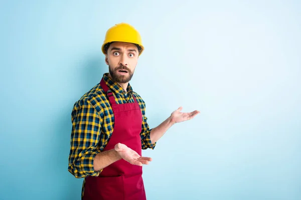 Confused workman in safety helmet showing shrug gesture on blue — Stock Photo
