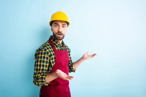 Verwirrter Arbeiter mit Schutzhelm zeigt achselzuckende Geste auf blauem Grund — Stockfoto