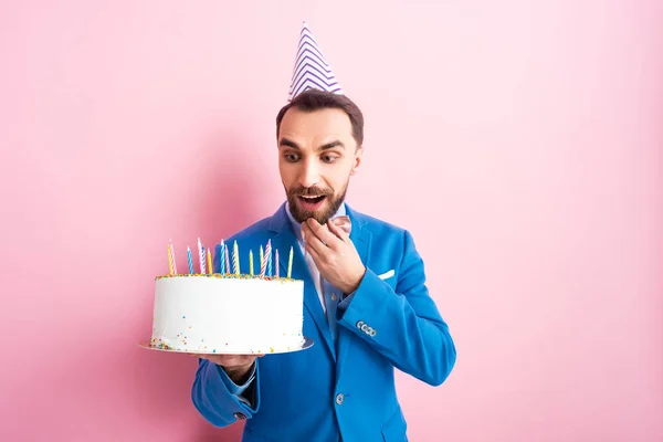 Здивований бородатий чоловік дивиться на торт на день народження на рожевому — стокове фото