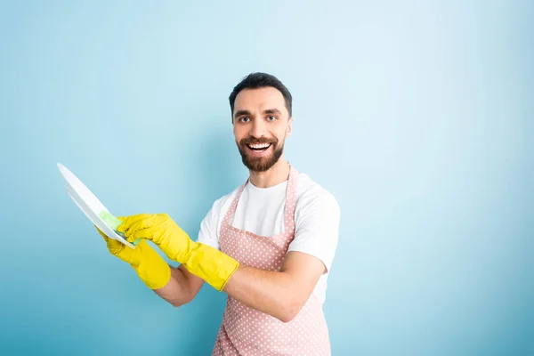 Fröhlicher bärtiger Mann wäscht Teller auf blauem Grund — Stockfoto