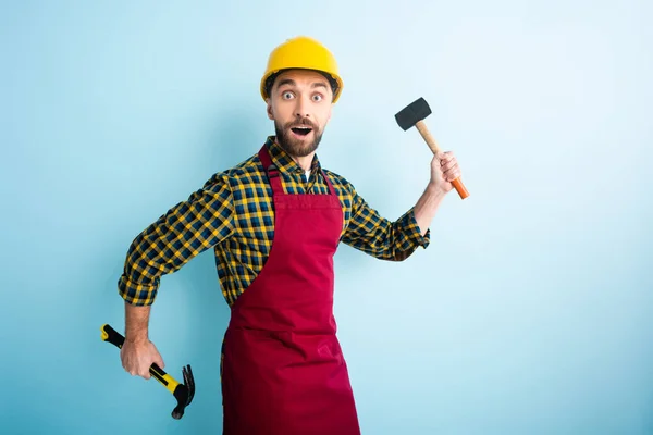 Surprised bearded man in safety helmet holding hammers on blue — Stock Photo