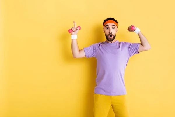 Homem barbudo surpreso apontando com o dedo enquanto segurava pequenos halteres no amarelo — Fotografia de Stock