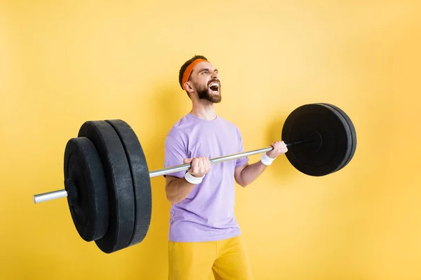 Емоційний бородатий спортсмен, що працює з барбелом на жовтому — стокове фото