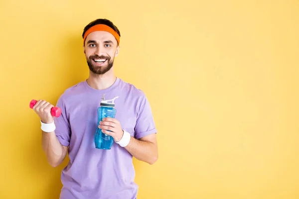 Bearded sportsman holding sports bottle and dumbbell on yellow — Stock Photo