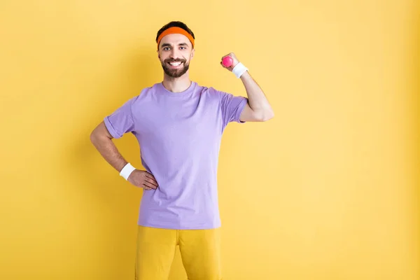 Веселий спортсмен тримає гантелі, стоячи з рукою на стегні на жовтому — стокове фото
