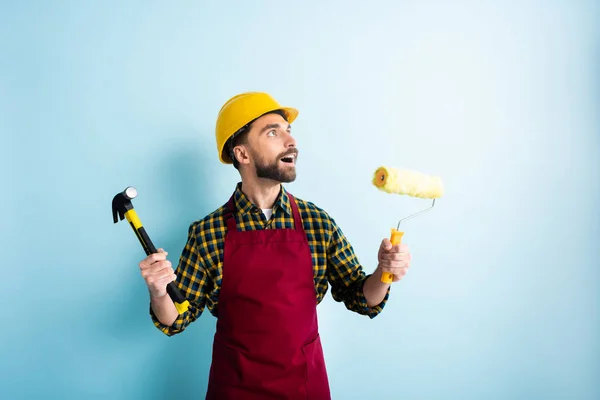 Ouvrier surpris dans le casque de sécurité tenant marteau et rouleau de peinture tout en regardant vers le haut sur bleu — Stock Photo