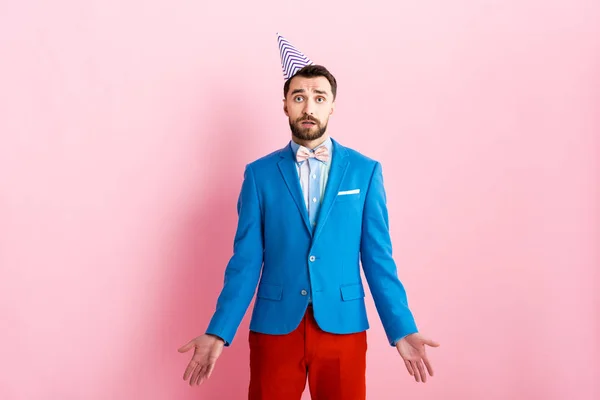 Verwirrter Geschäftsmann mit Partymütze zeigt Achselzucken-Geste auf rosa — Stockfoto