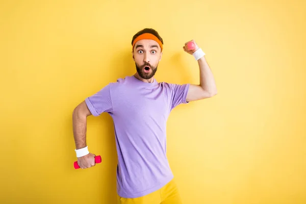 Shocked bearded man looking at camera while holding small dumbbells on yellow — Stock Photo