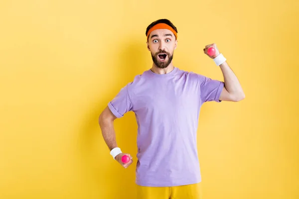 Sorprendido hombre barbudo mirando a la cámara mientras sostiene pequeñas mancuernas en amarillo - foto de stock