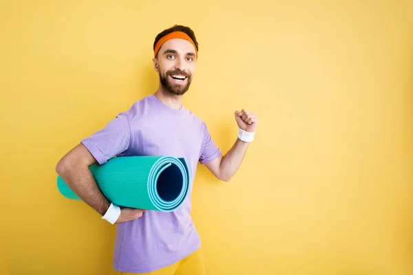 Sportif barbu joyeux tenant tapis de fitness sur jaune — Photo de stock