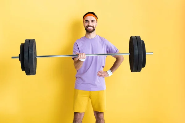 Cheerful sportsman standing with hand on hip and holding barbell on yellow — Stock Photo