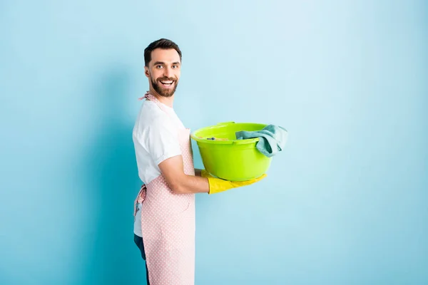 Positive bearded man in apron holding dirty laundry on blue — Stock Photo