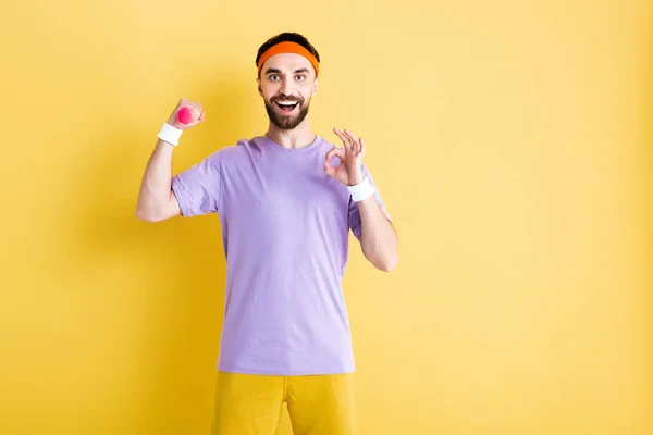Alegre deportista sosteniendo pequeña mancuerna y mostrando ok signo en amarillo — Stock Photo