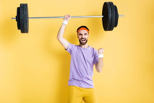 Excited sportsman celebrating while working out with barbell on yellow — Stock Photo