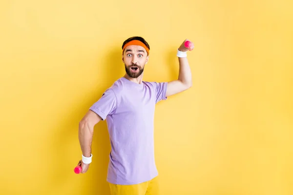 Surprised man looking at camera while holding small dumbbells on yellow — Stock Photo