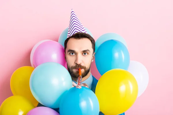 Bärtiger Mann mit Partymütze und Partygebläse im Mund in der Nähe von Luftballons auf rosa — Stockfoto