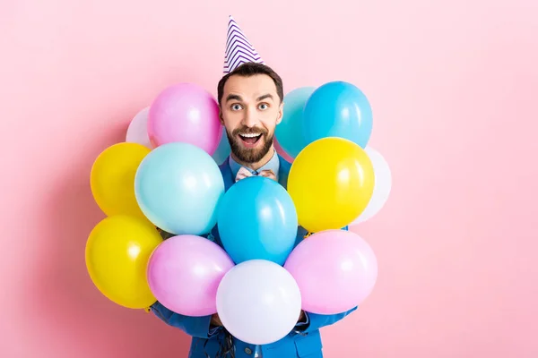 Homme barbu en chapeau de fête souriant près de ballons colorés sur rose — Photo de stock