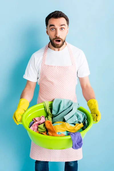 Shocked man in rubber gloves holding wash bowl with dirty laundry on blue — Stock Photo