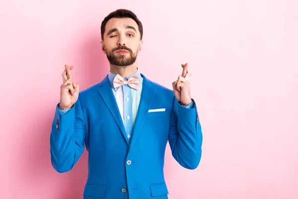 Hombre barbudo de traje con los dedos cruzados guiñando el ojo en rosa — Stock Photo