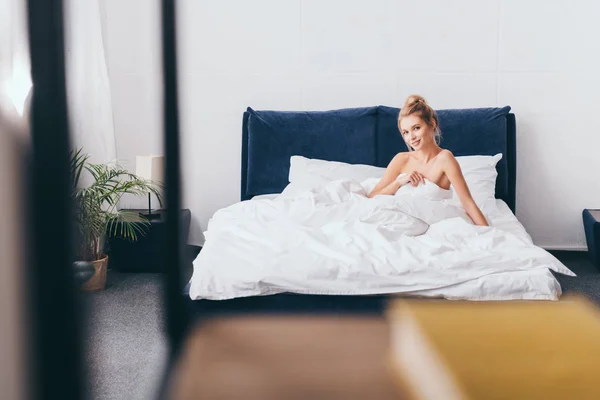 Mujer sonriente en sábanas sentadas en la cama por la mañana - foto de stock