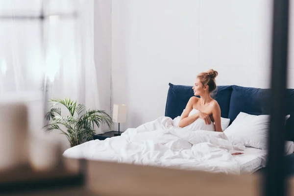 Beautiful happy woman in sheets sitting on bed and looking into window — Stock Photo