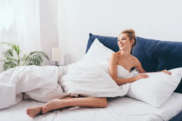 Schöne lächelnde Frau, die morgens in Laken auf dem Bett liegt — Stockfoto