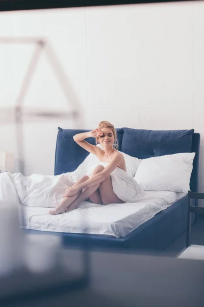 Enfoque selectivo de la hermosa mujer feliz en sábanas sentadas en la cama por la mañana - foto de stock