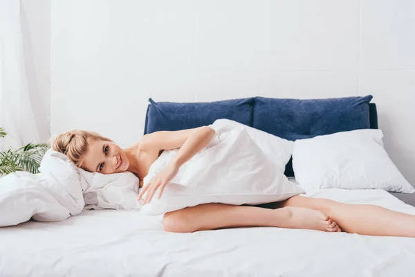 Femme souriante couchée dans des draps sur le lit le matin — Photo de stock