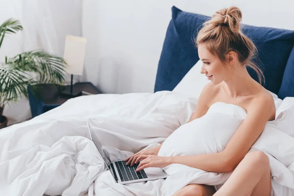 Mulher sorrindo usando laptop na cama com lençóis de manhã — Fotografia de Stock