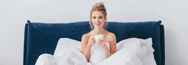 Panoramic shot of attractive happy woman with cup of coffee sitting in bed in morning — Stock Photo