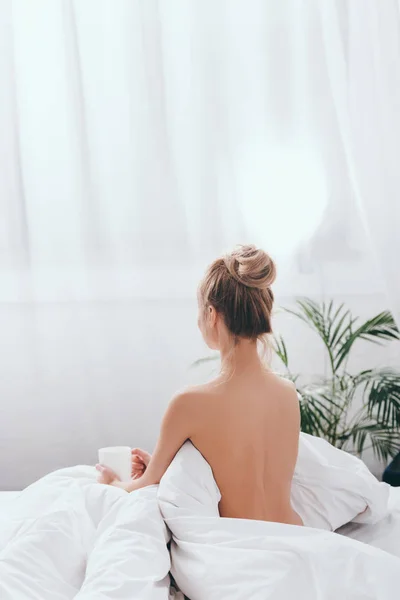 Rückansicht einer nackten Frau mit einer Tasse Kaffee, die morgens im Bett sitzt — Stockfoto